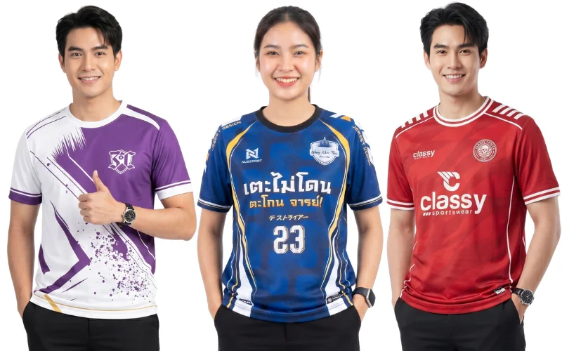 ตัวอย่างเสื้อกีฬาพิมพ์ลายและเสื้อทีมฟุตบอลสั่งทำ 3 รูปแบบ ซ้าย: เสื้อกีฬาพิมพ์ลายกราฟิกสีม่วงตัดขาวดีไซน์ทันสมัย กลาง: เสื้อฟุตบอลพิมพ์ลายสีน้ำเงินทองสกรีนข้อความกวนๆ "เตะไม่โดน ตะโกน จารย์!" เบอร์ 23 ขวา: เสื้อกีฬาคอกลมสีแดงลายคลาสสิกสไตล์สปอร์ต แสดงงานพิมพ์ซับลิเมชัน (Sublimation) สีสดคมชัดจากโรงงานผลิตเสื้อผ้า