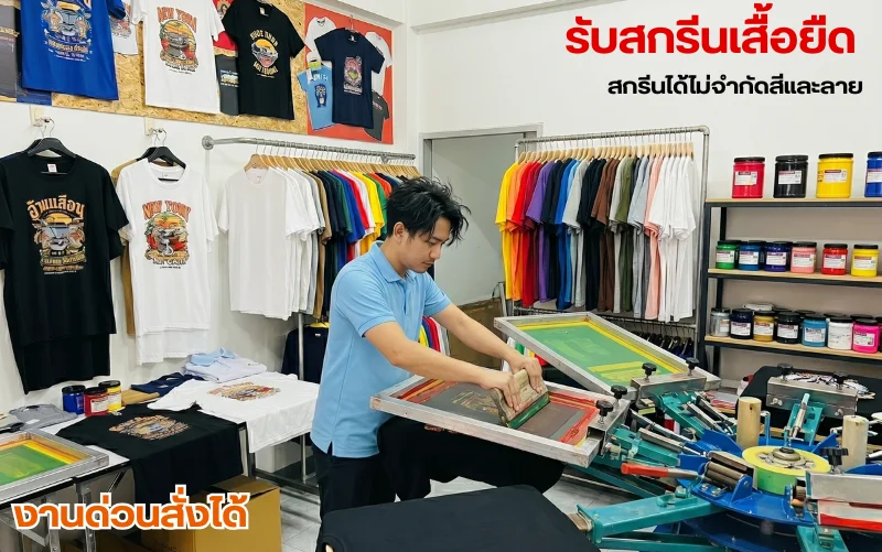 ภาพบรรยากาศในร้านหรือโรงงานสกรีนเสื้อผ้าที่สะอาดและเป็นระเบียบ มีพนักงานชายสวมเสื้อโปโลสีฟ้ากำลังตั้งใจใช้ไม้ปาดสกรีนทำงานร่วมกับเครื่องสกรีนเสื้อแบบตลับหมุนสีฟ้า (Carousel Screen Printing Machine) รอบ ๆ ห้องมีเสื้อยืดสีสันต่าง ๆ ทั้งเสื้อเปล่าและเสื้อที่สกรีนลายกราฟิกเสร็จแล้วแขวนโชว์บนราวและผนัง พร้อมชั้นวางกระปุกสีสกรีนจำนวนมาก โดยมีข้อความกราฟิกสีแดงระบุว่า "รับสกรีนเสื้อยืด สกรีนได้ไม่จำกัดสีและลาย" และมุมซ้ายล่างมีข้อความสีส้มขอบขาวระบุว่า "งานด่วนสั่งได้"

รายละเอียดเพิ่มเติมเชิงลึก:

จุดโฟกัสตรงกลาง: ช่างสกรีนชายเสื้อโปโลสีฟ้ากำลังโน้มตัวทำงานอยู่บนบล็อกสกรีนสี่เหลี่ยมขนาดใหญ่ ซึ่งติดตั้งอยู่บนโครงเครื่องสกรีนหมุนวงล้อสีฟ้า

ราวแขวนเสื้อด้านหลัง: ฝั่งขวาเป็นราวแขวนเสื้อยืดเปล่าหลากสีเรียงตามเฉดสี (แดง, เหลือง, น้ำเงิน, ดำ, เทา, ขาว) ส่วนฝั่งซ้ายเป็นราวแขวนเสื้อยืดสีขาวและเสื้อตัวอย่างที่พิมพ์ลายกราฟิกแนวสตรีท สไตล์วินเทจสีสันสดใส

ชั้นวางสีสกรีน: ด้านขวามีชั้นไม้ 3 ชั้นที่จัดวางกระปุกสีสำหรับงานสกรีนไว้แน่นขนัด มีฝาปิดหลากสี (แดง, ดำ, เหลือง, น้ำเงิน, เขียว, ชมพู) บ่งบอกถึงความพร้อมในการผสมสีและพิมพ์ลายที่หลากหลาย

การจัดวางสินค้าตัวอย่าง: บนโต๊ะด้านซ้ายมีเสื้อยืดที่สกรีนลายเสร็จแล้ววางเรียงกันเพื่อตรวจสอบความเรียบร้อย และมีบล็อกสกรีนเปล่าวางเตรียมพร้อมไว้ข้าง ๆ